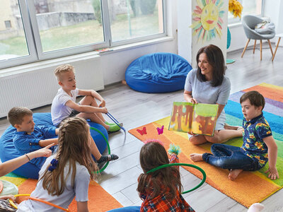 Eine Erzieherin sitzt lächelnd auf einer bunten Matte in einem hellen Raum und liest fünf Kindern aus einem Bilderbuch vor. Die Kinder sitzen im Kreis um sie herum, einige auf Sitzkissen, und hören aufmerksam zu. Ein Junge hält einen Hula-Reifen in der Hand. Im Hintergrund sind große Fenster mit Blick nach draußen, ein bunter Sitzsack, ein Stuhl und eine gemalte Sonne an der Wand zu sehen. Der Raum ist einladend und kindgerecht gestaltet.