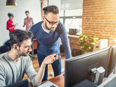 Zwei Personen im Vordergrund, eine sitzt und eine steht, und diskutieren, während sie auf einen Computerbildschirm schauen. Im Hintergrund unterhalten sich zwei weitere Personen in der Nähe eines Fensters. Die Umgebung scheint ein Büro mit Backsteinwänden und großen Fenstern zu sein.