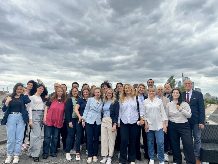Die Teilnehmenden stehen auf dem Dachgarten es Gebäudes für ein Gruppenfoto parat, im Hintergrund ist die Frankfurter Skyline mit Helaba und Commerzbank erkennbar. Am Himmel sind dunkle Gewitterwolken und den Damen weht der Wind durch die langen Haare.