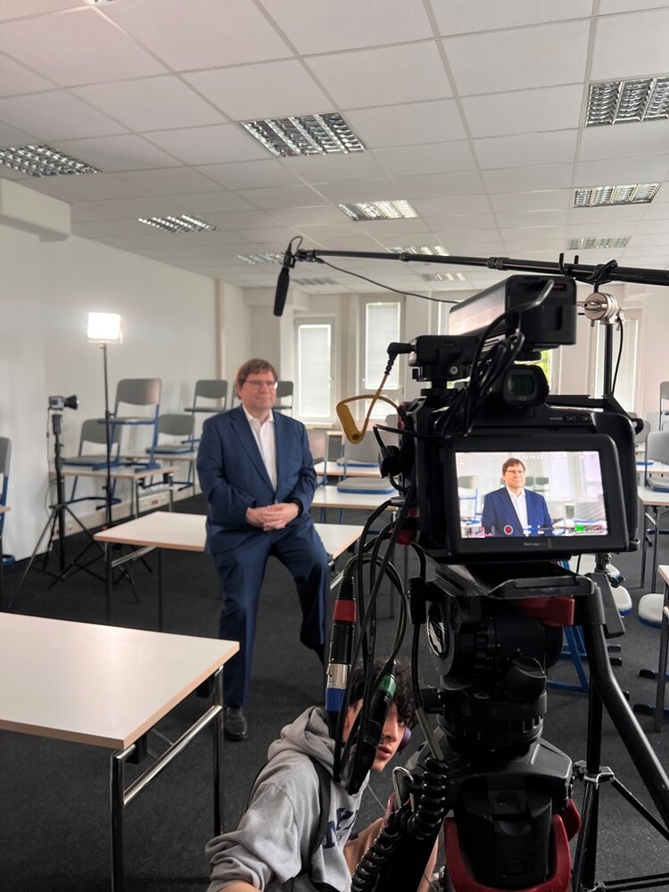 Ein Mann im Anzug sitzt auf einem Tisch in einem hellen Raum mit weißen Wänden und mehreren leeren Stühlen. Im Vordergrund steht eine professionelle Videokamera mit Mikrofon und Monitor, auf dem der Mann ebenfalls zu sehen ist. Ein Scheinwerfer beleuchtet die Szene. Im unteren Bildbereich ist eine Person mit Kapuze zu erkennen, die sich neben der Kamera befindet.