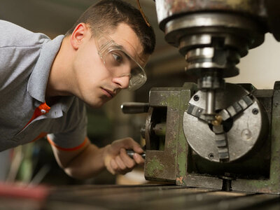 Eine Person bedient eine Metall-Drehmaschine. Das Gesicht der Person ist verschwommen, und sie trägt ein graues Poloshirt mit orangefarbenem Besatz am Ärmel. Die Person scheint die Maschine zu justieren oder zu inspizieren, die ein rotierendes Spannfutter hält, das ein Stück Material umfasst.