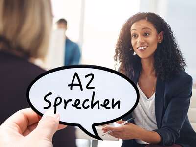 Eine lächelnde Person mit lockigen, dunklen Haaren und einer dunklen Blazerjacke über einem grauen Oberteil hält eine Sprechblase mit der Aufschrift 'A2 Sprechen' in den Händen. Im Hintergrund sind zwei weitere Personen unscharf zu erkennen, die sich unterhalten. Die Szene wirkt dynamisch und kommunikativ.