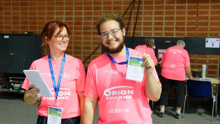 Gemeinsam im Einsatz für die PingPongParkinson-WM 2026 Zwei lächelnde Personen in pinken Volunteer-T-Shirts mit Namensschildern stehen in einer Halle; eine Person hält einen Ausweis in die Kamera.