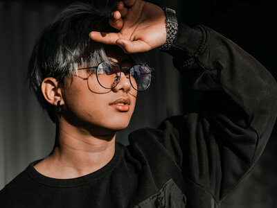 Eine Person mit kurzen, dunkelgrauen Haaren und einer Brille steht im Profil. Sie trägt ein schwarzes Sweatshirt und hat eine Hand über den Augen, als würde sie die Sonne abschirmen. Die Person trägt eine Armbanduhr und Ohrringe.