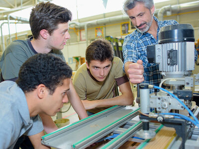 Vier Personen stehen um eine Maschine in einer Werkstatt- oder Industrieumgebung. Die Maschine besteht aus Metallschienen und verschiedenen mechanischen Komponenten, möglicherweise Teil eines Fertigungs- oder Technikprozesses. Eine Person bedient die Maschine, während die anderen aufmerksam zusehen.