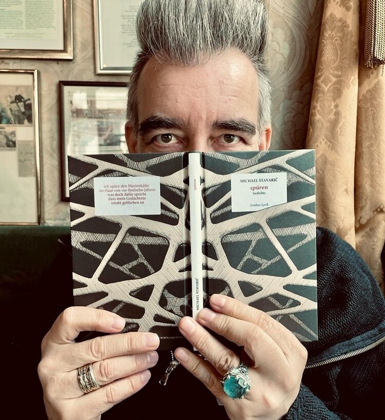 Schriftsteller Michael Stavaric trägt zwei große Ringe, einen mit opulentem grünem Edelstein, an den Fingern, der hält seinen neuen Gedichtband "spüren" bis zu den Augen vors Gesicht und posiert, mit seinen grauen, hübsch nach oben frisierten Haaren, für ein Foto.
