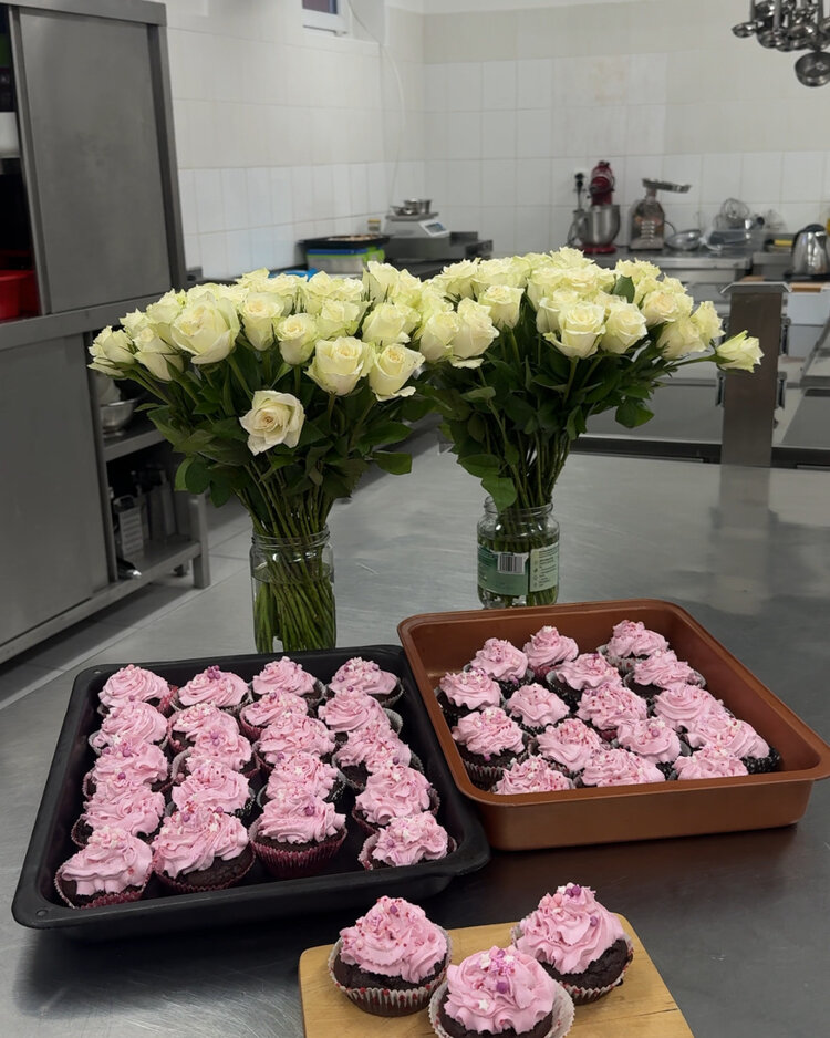 Auf einem Tisch stehen zwei große Trays mit rosa verzierten Cupcakes. Dahinter sind zwei Vasen mit großen Sträußen weißer Rosen zu sehen. Im Hintergrund befindet sich eine Küche mit Arbeitsflächen und Geräten.