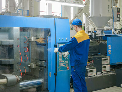 Ein Arbeiter bedient ein Bedienfeld an einer großen blauen Industriemaschine, die wahrscheinlich für die Herstellung oder Verarbeitung verwendet wird. Der Arbeiter trägt blau-gelbe Arbeitskleidung, einschließlich einer Mütze und Handschuhen. Die Maschine hat verschiedene Knöpfe, Schalter und Anzeigen am Bedienfeld. Durch einen transparenten Abschnitt der Maschine sind auch rote Kabel sichtbar. Im Hintergrund sind weitere industrielle Geräte und Maschinen zu sehen.