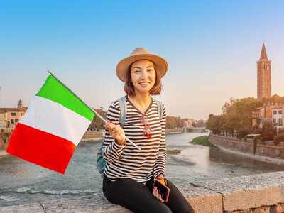 Eine lächelnde Frau sitzt auf einer Steinmauer und hält eine italienische Flagge in der Hand. Sie trägt einen Hut, ein gestreiftes Langarmshirt und eine Sonnenbrille auf dem Kopf. Im Hintergrund fließt ein Fluss, und es ist ein sonniger Tag mit einem klaren, blauen Himmel. Ein historisches Gebäude mit einem Turm ist ebenfalls im Hintergrund zu sehen.