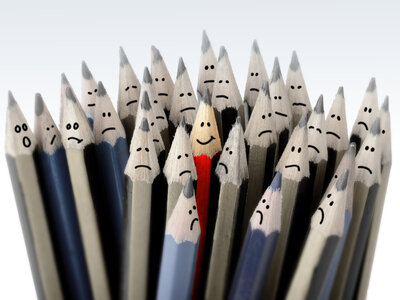 Eine Gruppe von Bleistiften, die alle traurige Gesichter haben, außer einem Bleistift in der Mitte, der ein lächelndes Gesicht hat. Der lächelnde Bleistift ist rot und steht im Vordergrund, während die anderen Bleistifte in verschiedenen Grau- und Blautönen gehalten sind.