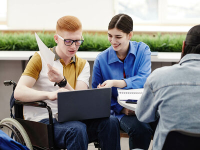 Drei Personen sitzen zusammen an einem Tisch. Ein junger Mann mit roten Haaren und einer Brille sitzt im Rollstuhl und hält ein Blatt Papier. Neben ihm sitzt eine lächelnde Frau mit einem blauen Hemd, die auf einen Laptop schaut. Eine weitere Person mit langem, dunklem Haar sitzt mit dem Rücken zur Kamera und scheint ebenfalls an der Diskussion teilzunehmen. Im Hintergrund sind Pflanzen zu sehen.