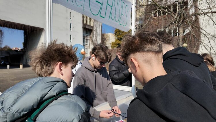 Global Money Week 2026 an der Euroskola Praha Eine Gruppe junger Menschen stehen an einem Stand und beobachtet einen jungen Mann mit Sonnenbrille, der mit einer Zange hantiert. Im Hintergrund hängt ein Banner, auf der man das Wort 'Glasses' erkennen kann.