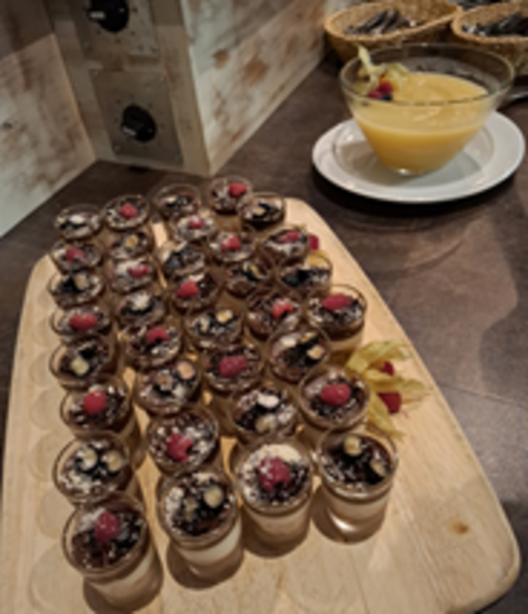 Holzbrett mit vielen kleinen Dessertgläsern, gefüllt mit geschichteter Creme und Schokostreuseln, jeweils mit Beeren und Nüssen garniert; dahinter eine Schale mit gelber Sauce.