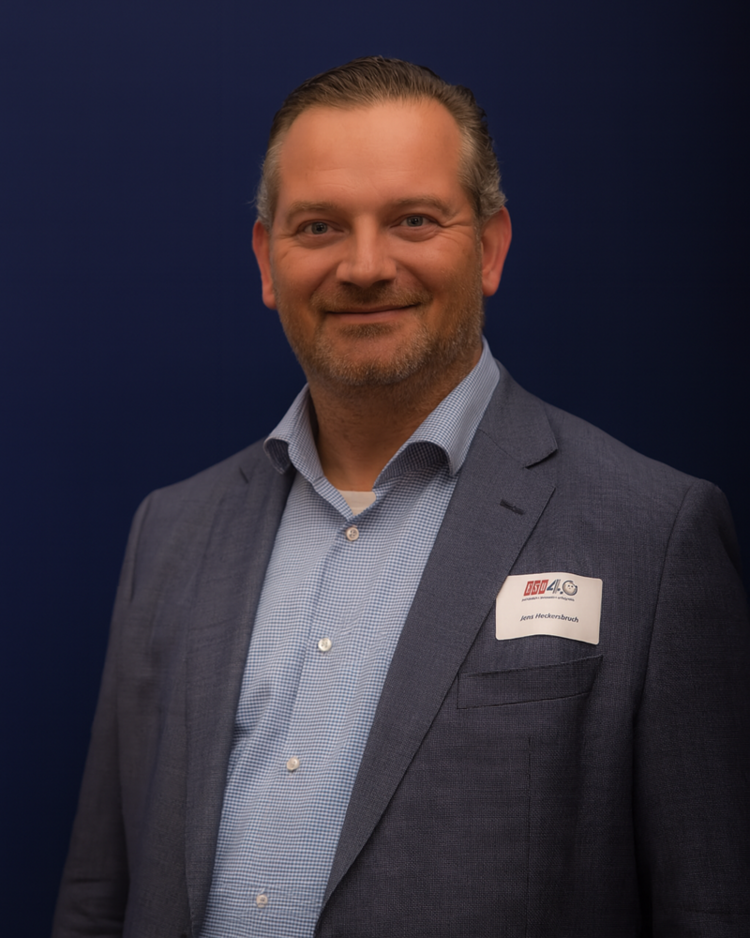 Mitarbeiter Porträt-Foto von Jens Heckersbruch, der ein weiß-blau karriertes Hemd und ein mausgrau-blaues Sacko trägt und freundlich in die Kamera lächelt. 