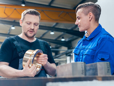 Zwei Personen in einer Werkstatt oder industriellen Umgebung. Eine Person hält ein metallisches zylindrisches Objekt mit beiden Händen, während die andere Person daneben steht und zuschaut. Im Hintergrund sind industrielle Geräte und Strukturen wie Balken und Lichter zu sehen. Die Gesichter der Personen sind aus Datenschutzgründen verschwommen.