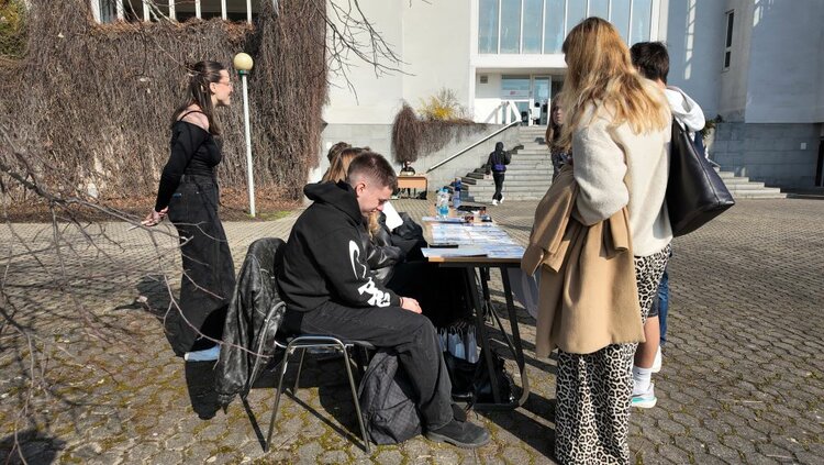 Global Money Week 2026 an der Euroskola Praha Mehrere Personen sitzen nebeneinander an einem Tisch im Freien, auf dem mehrere Dokumente ausgelegt sind. Andere Personen davor. Im Hintergrund sind ein Gebäude und ein Baum zu sehen.