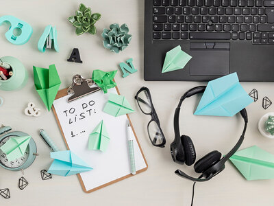 Ein organisierter Schreibtisch mit verschiedenen Gegenständen, darunter ein Clipboard mit einer To-Do-Liste, auf der '1...' geschrieben steht. Auf dem Tisch befinden sich auch eine Tastatur, ein Headset, eine Brille, ein Stift, ein Wecker, mehrere Origami-Figuren in Grün und Blau, kleine Sukkulenten in Töpfen, ein Klebebandabroller, ein Tacker und verschiedene Büroklammern. Die Gegenstände sind auf einem hellen Hintergrund angeordnet.