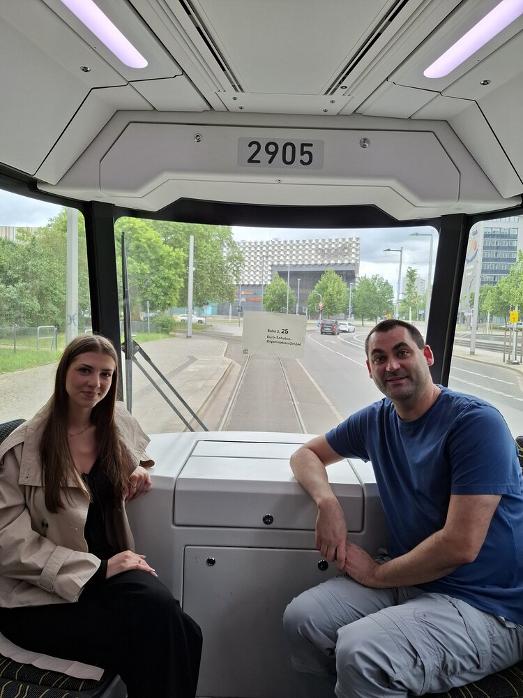 Im Inneren einer Straßenbahn sitzen links eine Frau und rechts ein Mann vor dem Fenster am Ende der Bahn und schauen in die Kamera.
