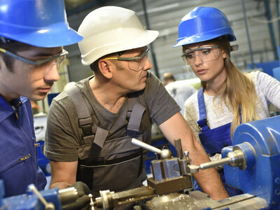 Drei Personen in einer industriellen Umgebung arbeiten an einer Maschine. Die Person in der Mitte, ein Mann mit einem weißen Helm und einer Schutzbrille, scheint Anweisungen zu geben oder eine Erklärung abzugeben. Die Person links, ein Mann mit einem blauen Helm und einer Schutzbrille, beobachtet aufmerksam. Die Person rechts, eine Frau mit einem blauen Helm und einer Schutzbrille, bedient die Maschine. Alle tragen Arbeitskleidung und Sicherheitsausrüstung.