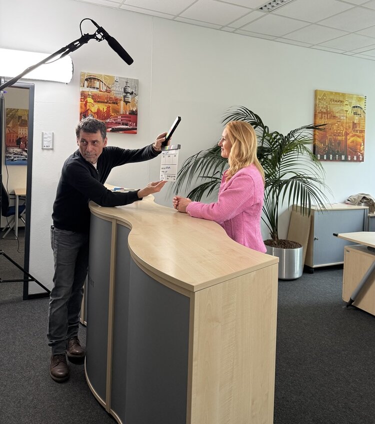 Eine Frau mit langen, blonden Haaren und rosa Blazer steht an einem Empfangstresen aus hellem Holz mit grauen Fronten. Ein Mann hält eine Filmklappe und ein Mikrofon an einer Tonangel über sie. Im Hintergrund stehen ein großer, grüner Blumentopf und zwei farbige Stadtansichten an der Wand. Die Szene spielt in einem modern eingerichteten Büro mit Teppichboden und abgehängter Decke.