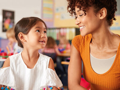 Auf dem Bild sind zwei Personen im Klassenzimmer zu sehen. Die Person links trägt eine weiße Bluse mit bunten Pompons an den Ärmeln, während die Person rechts ein orangefarbenes Oberteil und lockiges Haar hat. Im Hintergrund sind Kinderzeichnungen, pädagogische Poster und andere Lernmaterialien an den Wänden zu erkennen.