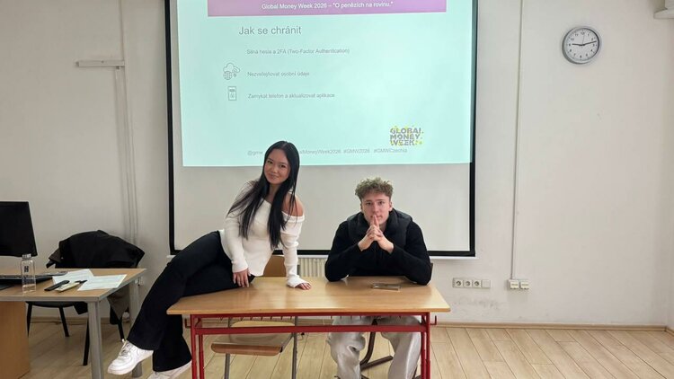 Global Money Week 2026 an der Euroskola Praha Ein junger Mann sitzt mit gefalteten Händen auf einem Stuhl an einem Tisch vor einer Leinwand in einem Seminarraum. Eine junge Frau mit langem Haar sitzt lässig daneben auf dem Tisch und stützt sich mit einem Arm darauf ab. Auf der Leinwand steht 'Jak se chránit' und es geht um das Thema 'Global Money Week 2026 – O penězích na rovinu'. Die beiden Personen posieren lächelnd für das Foto.