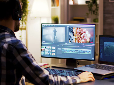 Ein Mensch mit Kopfhörern sitzt an einem Schreibtisch und bearbeitet Videomaterial auf einem großen Bildschirm. Auf dem Bildschirm sind verschiedene Videoclips und eine Audio-Timeline zu sehen. Ein Laptop steht daneben auf dem Tisch.