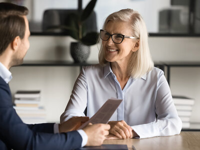 Eine ältere Frau mit kurzen, hellen Haaren und einer Brille sitzt an einem Tisch in einem modernen Büro. Sie trägt ein helles, langärmliges Hemd und hält ein Dokument in den Händen. Ihr gegenüber sitzt ein Mann in einem dunklen Anzug, der ihr das Dokument überreicht. Beide wirken konzentriert und professionell. Im Hintergrund sind Regale mit Ordnern und eine große Zimmerpflanze zu sehen. Die Atmosphäre ist seriös und geschäftlich, was auf ein Vorstellungsgespräch oder eine wichtige Besprechung hindeutet.