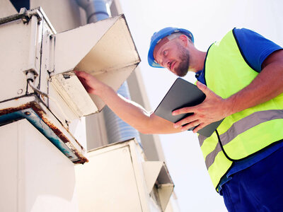 Ein Techniker mit blauer Schutzbrille und neon-gelber Warnweste arbeitet an einer Klimaanlage oder Lüftungsanlage an einer Außenwand. Er hält ein Tablet in der Hand und berührt gleichzeitig die Anlage, möglicherweise um Daten abzulesen oder Einstellungen vorzunehmen. Der Hintergrund zeigt einen klaren Himmel.