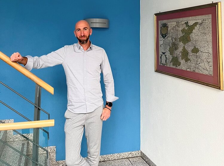 Mitarbeiter steht im Treppenhaus, vor blauer Wand, mit einem Arm an das Geländer gelehnt und posiert lächelnd für ein Foto.