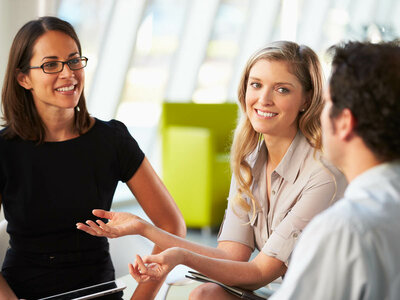 Drei Personen in einem Büro, zwei Frauen und ein Mann, sind in ein Gespräch vertieft. Die Frau links trägt ein schwarzes Oberteil und eine Brille, die Frau in der Mitte trägt eine beige Bluse, und der Mann rechts ist von hinten zu sehen.