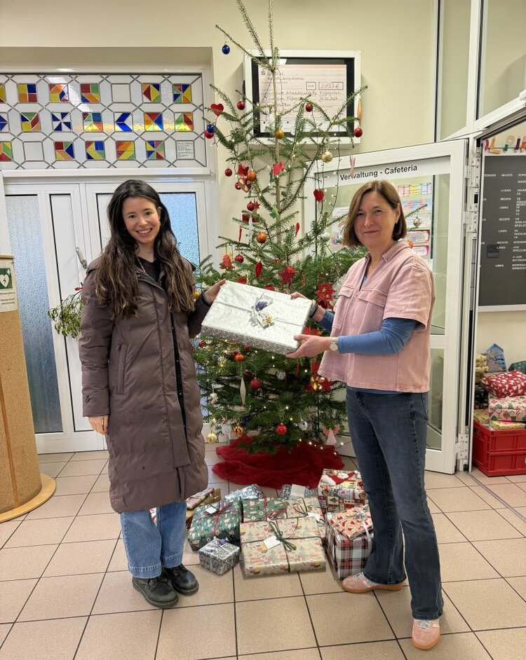 Zwei lächelnde Frauen stehen vor einem geschmückten Weihnachtsbaum in einer Eingangshalle. Die Frau links trägt einen langen, dunklen Mantel und blaue Jeans, die Frau rechts trägt ein rosafarbenes Oberteil und eine blaue Bluse. Beide halten gemeinsam ein großes, weiß-silbern verpacktes Geschenk. Um den Baum herum liegen zahlreiche bunt verpackte Geschenke auf dem Boden. Im Hintergrund sind farbenfrohe Glasfenster und ein schwarzes Tafelmenü an der Wand zu sehen.
