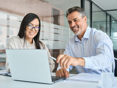 Eine Frau und ein Mann sitzen lächelnd an einem Tisch in einem modernen Büro. Die Frau trägt eine Brille und ein helles, gestreiftes Oberteil, während sie auf den Laptop-Bildschirm schaut. Der Mann, der ein hellblaues Hemd trägt, zeigt auf den Bildschirm und hält einen Stift in der Hand. Beide wirken konzentriert und zufrieden. Auf dem Tisch liegen ein Notizblock und ein Glas mit Wasser. Im Hintergrund sind Glaswände und ein offener Bürobereich zu sehen.