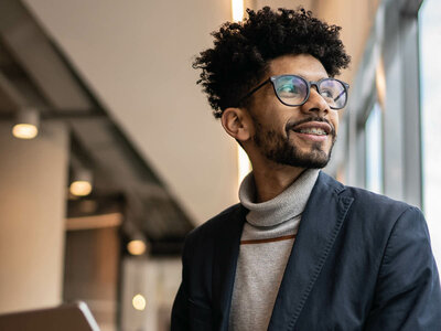 Ein lächelnder Mann mit Afro-Frisur und Brille, der einen grauen Rollkragenpulli und ein dunkles Sakko trägt. Er steht in einem modernen Innenraum, möglicherweise einem Büro oder Café, mit einem leicht unscharfen Hintergrund, der Regale und Beleuchtung zeigt.