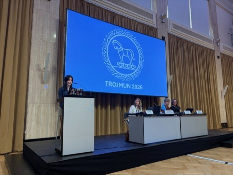 Eine Person hält eine Rede an einem Rednerpult auf einer Bühne. Im Hintergrund zeigt eine große Leinwand das Logo und den Schriftzug „TROJMUN 2026“. Drei Personen sitzen an einem Tisch rechts auf der Bühne.