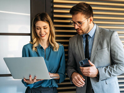 Zwei Personen stehen in einem modernen Büro und arbeiten gemeinsam. Die Frau links trägt eine blaue Bluse und hält einen silbernen Laptop in den Händen. Sie lächelt und wirkt konzentriert. Der Mann rechts trägt einen grauen Anzug, ein blaues Hemd und eine Brille. Er hält ein Smartphone in der Hand und blickt darauf. Im Hintergrund sind eine weiße Tafel und eine Holzvertäfelung zu sehen. Die Atmosphäre ist professionell, kooperativ und zielorientiert.