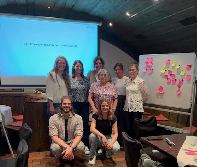 Acht Teammitglieder des Funktionsbereichs Business Development stehen und sitzen gemeinsam lächelnd vor einer Leinwand, auf der „DANKE an euch allen für den tollen Einstieg!“ steht. Rechts im Bild befindet sich ein Whiteboard mit bunten Post-its. Die Atmosphäre wirkt freundlich und kooperativ.