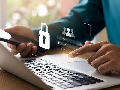 Eine Person arbeitet an einem Laptop und hält gleichzeitig ein Smartphone in der Hand. Auf dem Laptop-Bildschirm ist ein Login-Fenster zu sehen, das Benutzername und Passwort abfragt. Über dem Laptop schwebt ein Symbol eines Vorhängeschlosses, das Sicherheit oder Datenschutz symbolisiert, sowie ein Benutzersymbol, das auf den Login-Prozess hinweist.
