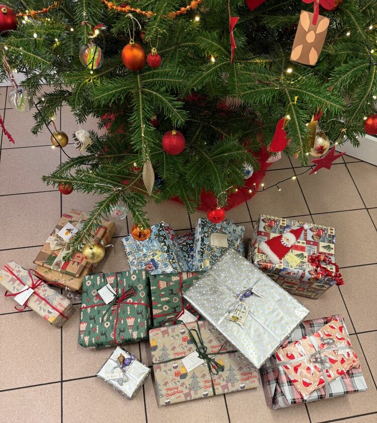 Ein Ausschnitt eines geschmückten Weihnachtsbaums zeigt grüne Zweige, die mit roten und goldenen Kugeln, Lametta und kleinen Figuren dekoriert sind. Unter dem Baum liegen zahlreiche bunt verpackte Geschenkpäckchen in verschiedenen Mustern und Größen auf dem gefliesten Boden. Die Geschenke sind mit Schleifen und Geschenkbändern verziert.