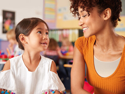 Auf dem Bild sind zwei Personen im Klassenzimmer zu sehen. Die Person links trägt eine weiße Bluse mit bunten Pompons an den Ärmeln, während die Person rechts ein orangefarbenes Oberteil und lockiges Haar hat. Im Hintergrund sind Kinderzeichnungen, pädagogische Poster und andere Lernmaterialien an den Wänden zu erkennen.