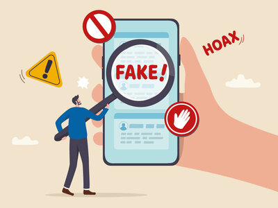 Eine Illustration zeigt eine große Hand, die ein Smartphone hält. Auf dem Smartphone-Bildschirm ist das Wort 'FAKE!' durch eine Lupe vergrößert zu sehen. Um das Smartphone herum befinden sich verschiedene Warnsymbole: ein rotes Verbotsschild, ein gelbes Ausrufezeichen in einem Dreieck und ein rotes Hand-Stopp-Schild. Ein Mann steht neben der Hand und deutet auf das Smartphone. Im Hintergrund ist das Wort 'HOAX' zu sehen.