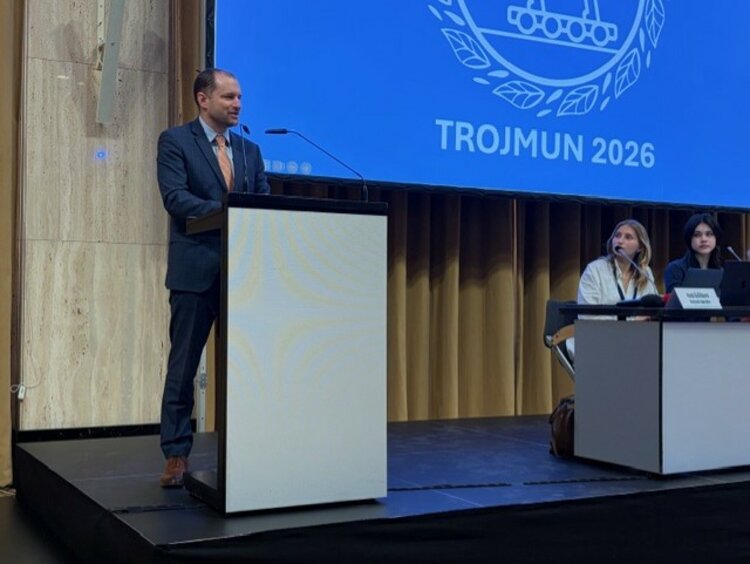 Ein Mann in einem dunkelblauen Anzug und lachsfarbener Krawatte spricht an einem Rednerpult auf einer Bühne. Im Hintergrund ist eine Leinwand mit dem Logo und dem Schriftzug „TROJMUN 2026“ zu sehen. Zwei Personen sitzen an einem Tisch rechts auf der Bühne.
