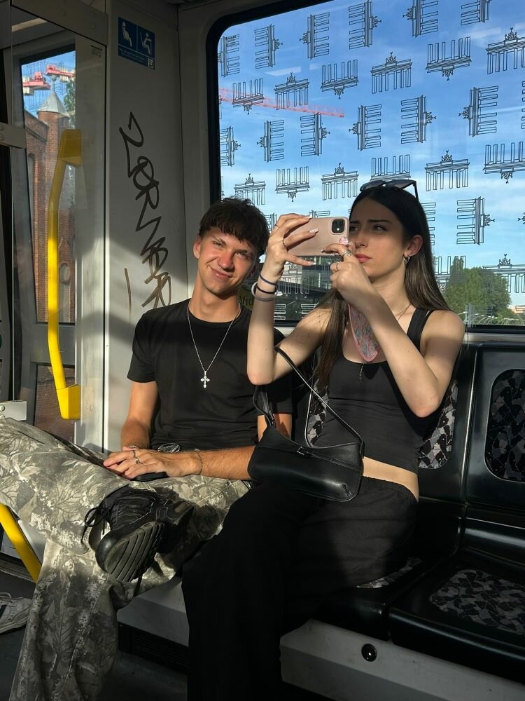 Klassenfahrt der Euroskola Prag nach Berlin Zwei Jugendliche sitzen nebeneinander in einem Straßenbahnabteil. Die Person links lächelt in die Kamera, während die Person rechts ein Handy in der Hand hält und ein Selfie macht. Im Hintergrund sind Fenster mit Motiven des Brandenburger Tors zu sehen.