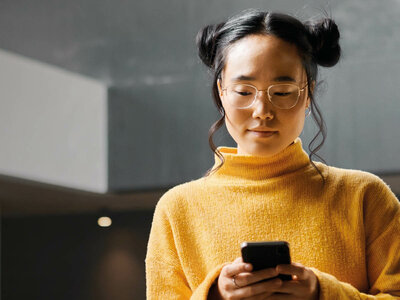 Eine Person mit Brille und zwei Zöpfen, die zu einem hohen Rollkragen-Pullover in Gelb trägt. Sie hält ein Smartphone in den Händen und scheint auf den Bildschirm zu schauen. Der Hintergrund ist unscharf, was darauf hindeutet, dass sie sich in einem Innenraum befindet.