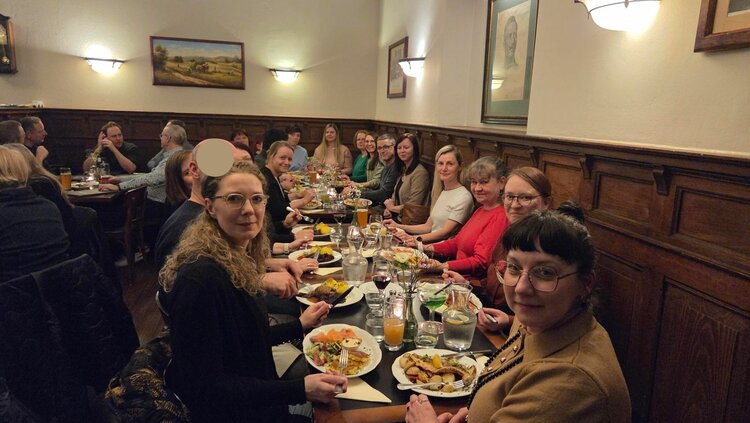 Große Gruppe sitzt an einer langen Tafel in einem gemütlichen Restaurant und blickt in die Kamera; auf dem Tisch stehen Teller mit Essen, Gläser und Kerzen.