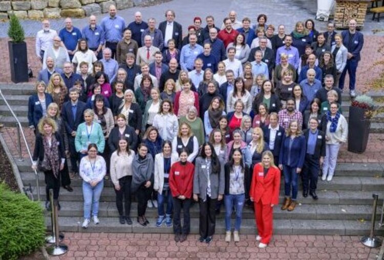 Große Gruppe mit Namensschildern steht in mehreren Reihen auf einer Außentreppe und blickt zur Kamera.