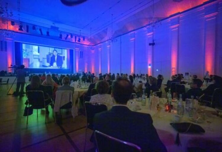 Gäste sitzen an festlich gedeckten Tischen im blau und rosa beleuchteten Saal und verfolgen ein Video auf einer großen Leinwand.