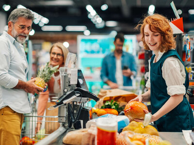 Eine junge Frau mit rotem Haar und einer Schürze steht an einer Supermarktkasse und scannt Artikel. Ein älterer Mann mit Brille und einem weißen Hemd legt eine Ananas auf das Förderband. Im Hintergrund sind weitere Kunden zu sehen, darunter eine Frau mit blonden Haaren und ein Mann mit einem blauen Hemd, der auf sein Smartphone schaut. Die Kasse ist mit verschiedenen Produkten wie Brot und Getränken gefüllt.
