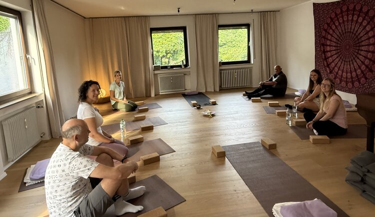 In einem Yogastudio haben sich es sechs Teilnehmende des Kurses auf ihren Matten bequem gemacht. An der Wand hängt ein weinrotes Tuch mit einem Mandala. Auf und neben den Matten liegen Klötze, Decken und Kissen. Der Kurs kann gleich beginnen.