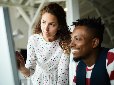 Eine junge Frau mit lockigen Haaren und einem weißen, gepunkteten Kleid steht lächelnd neben einem Mann, der eine rote Brille und ein rot-weißes Oberteil trägt. Beide wirken entspannt und freundlich. Die Frau deutet mit der Hand auf etwas außerhalb des Bildes, während der Mann sie lächelnd ansieht. Der Hintergrund zeigt einen hellen, modernen Innenraum, möglicherweise ein Büro oder ein öffentlicher Raum.